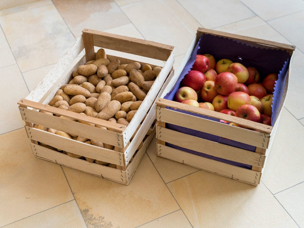 Eine Kiste mit Äpfeln und eine Kiste mit Kartoffeln stehen auf einem mit cremefarbenen Bodenplatten gepflasterten Boden